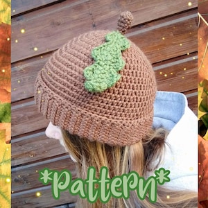Peut inclure: Un bonnet en crochet de couleur marron en forme de gland avec une feuille en crochet verte sur le dessus. Le bonnet est porté par une personne aux cheveux longs et bruns. Le texte "*Pattern*" est écrit en lettres vertes sur l'image.