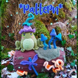 May include: Crochet pattern for a whimsical frog sitting on a log with mushrooms and a dragonfly. The frog is wearing a purple hat and has green eyes. The log is brown and has a green moss texture. The mushrooms are blue and the dragonfly is purple and blue.