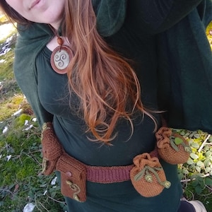 K&ouml;nnte beinhalten: Eine Frau tr&auml;gt ein dunkelgr&uuml;nes Kleid mit einem braunen geh&auml;kelten G&uuml;rtel und mehreren geh&auml;kelten Beuteln. Sie tr&auml;gt eine Halskette mit einem braunen Anh&auml;nger, der ein dreifaches Spiralmuster zeigt.