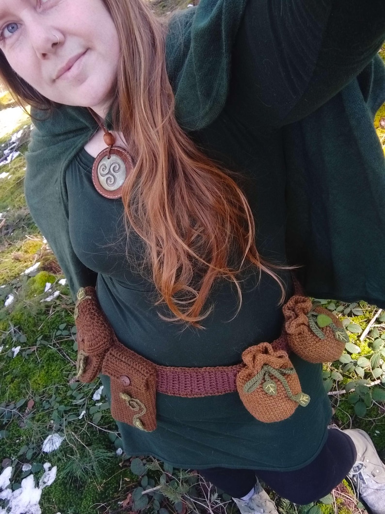 K&ouml;nnte beinhalten: Eine Frau tr&auml;gt ein dunkelgr&uuml;nes Kleid mit einem braunen geh&auml;kelten G&uuml;rtel und mehreren geh&auml;kelten Beuteln. Sie tr&auml;gt eine Halskette mit einem braunen Anh&auml;nger, der ein dreifaches Spiralmuster zeigt.