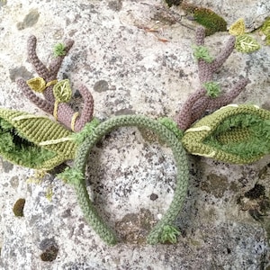 May include: A handcrafted, green and brown crochet headband with deer antlers and leaf accents. The antlers are a muted brown, while the leaves and headband are various shades of green. The design evokes a woodland or nature theme.