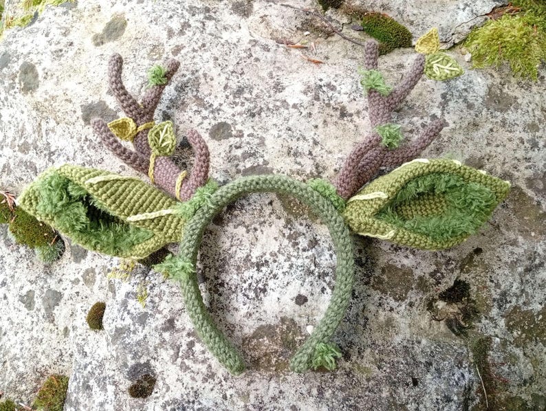 May include: A handcrafted, green and brown crochet headband with deer antlers and leaf accents. The antlers are a muted brown, while the leaves and headband are various shades of green. The design evokes a woodland or nature theme.