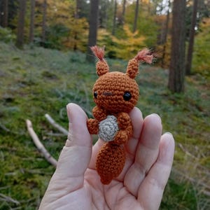 May include: A handmade crochet squirrel figurine in shades of brown and gray. The squirrel has small black eyes, a fluffy tail, and tufts of hair on its ears. The figurine is held in a hand against a blurred forest background.