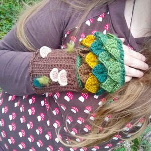 Puede incluir: Guantes de mu&ntilde;eca de ganchillo hechos a mano en marr&oacute;n, verde y amarillo. Los guantes de mu&ntilde;eca presentan un dise&ntilde;o de tronco de &aacute;rbol con acentos de seta y un patr&oacute;n de hojas. Ideales para oto&ntilde;o o invierno.