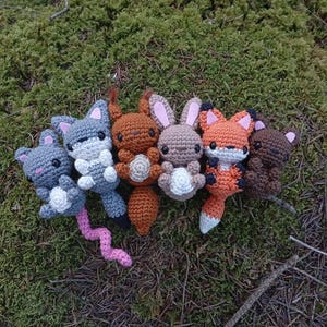 May include: A collection of six crocheted animal toys, including a gray mouse with a pink tail, a gray fox, a brown bear, and two brown rabbits. The toys are arranged on a bed of green moss and twigs.