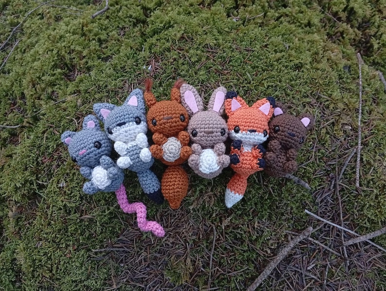 May include: A collection of six crocheted animal toys, including a gray mouse with a pink tail, a gray fox, a brown bear, and two brown rabbits. The toys are arranged on a bed of green moss and twigs.