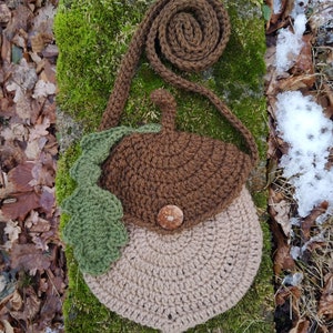 May include: A brown and beige crocheted acorn-shaped purse with a green crocheted leaf detail and a brown button. The purse has a brown crocheted strap.