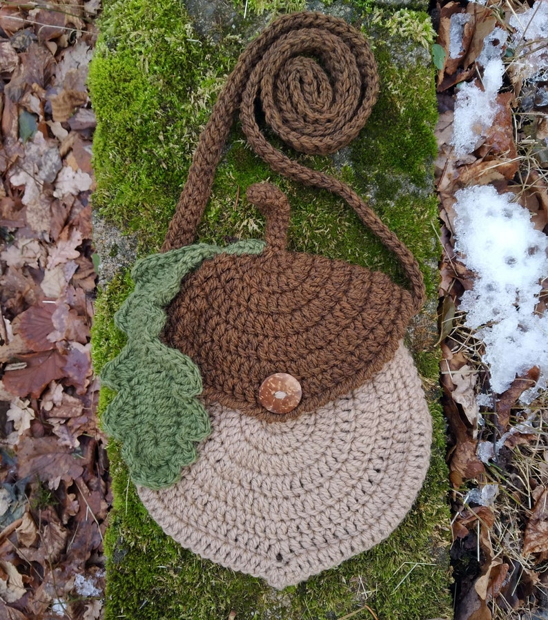 May include: A brown and beige crocheted acorn-shaped purse with a green crocheted leaf detail and a brown button. The purse has a brown crocheted strap.