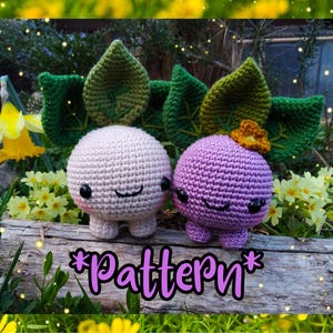 May include: Two crocheted amigurumi toys shaped like turnips with green leaves. The turnip on the left is white with a pink blush and the turnip on the right is purple with a yellow flower on top. The text "*Pattern*" is written in pink letters on a wooden surface.