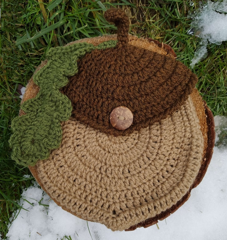 May include: A crocheted acorn hat with a brown cap, a tan bottom, and a green leaf. The hat has a wooden button.