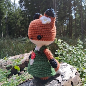 May include: A crocheted fox doll wearing a green dress with a red flower and a white flower on its head. The doll is sitting on a log in a forest setting.