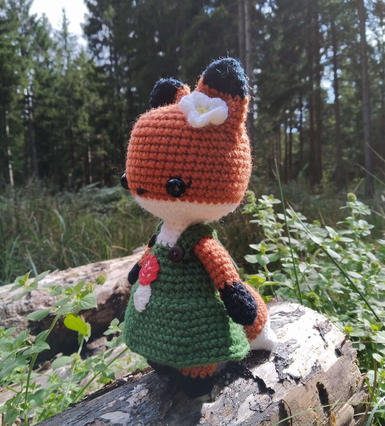 May include: A crocheted fox doll wearing a green dress with a red flower and a white flower on its head. The doll is sitting on a log in a forest setting.