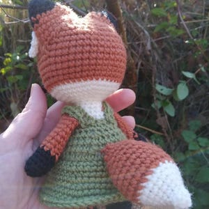 May include: A crocheted fox doll wearing a green dress. The fox has a white belly and a white tip on its tail. The doll is held in a hand.