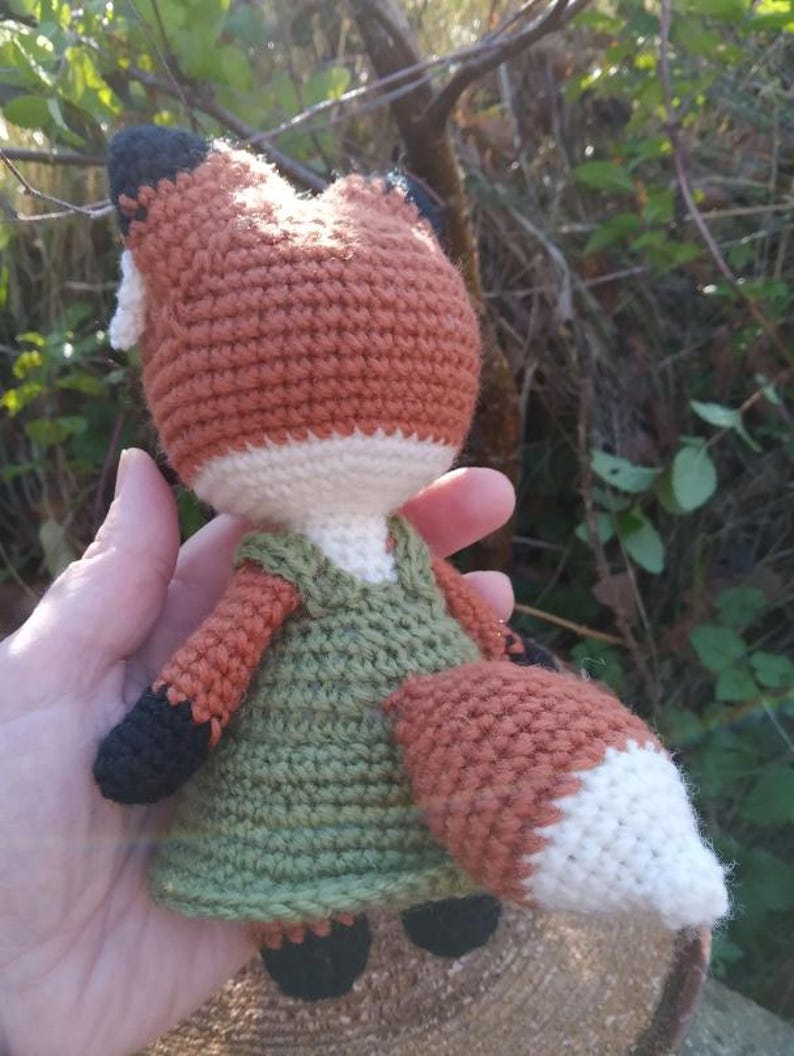 May include: A crocheted fox doll wearing a green dress. The fox has a white belly and a white tip on its tail. The doll is held in a hand.