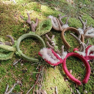 May include: A collection of handmade headbands with deer antler designs. The headbands are in various colors, including green, brown, and red. The antlers are made of a textured yarn and the ears are lined with faux fur.