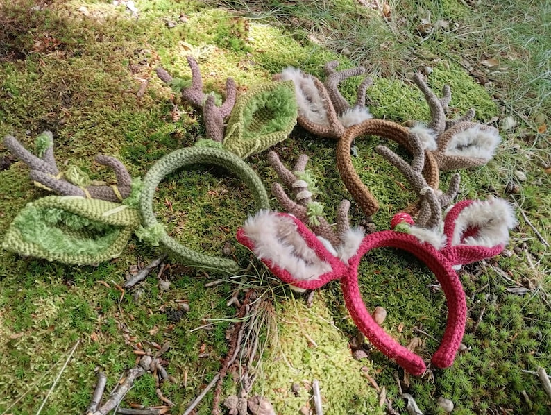 May include: A collection of handmade headbands with deer antler designs. The headbands are in various colors, including green, brown, and red. The antlers are made of a textured yarn and the ears are lined with faux fur.