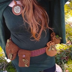 K&ouml;nnte beinhalten: Eine Frau tr&auml;gt einen gr&uuml;nen Kapuzenumhang und einen braunen geh&auml;kelten G&uuml;rtel mit mehreren Taschen. Der G&uuml;rtel hat einen braunen Knopf und gr&uuml;ne Blattembroidery. Sie tr&auml;gt auch eine braune Lederkette mit einem silbernen keltischen Knotenanh&auml;nger.