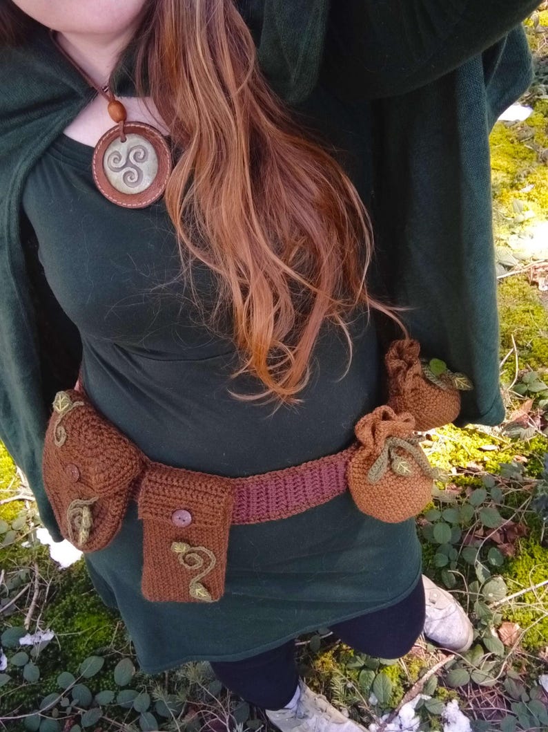 K&ouml;nnte beinhalten: Eine Frau tr&auml;gt einen gr&uuml;nen Kapuzenumhang und einen braunen geh&auml;kelten G&uuml;rtel mit mehreren Taschen. Der G&uuml;rtel hat einen braunen Knopf und gr&uuml;ne Blattembroidery. Sie tr&auml;gt auch eine braune Lederkette mit einem silbernen keltischen Knotenanh&auml;nger.
