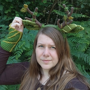 May include: A handmade crochet headband with deer antlers and matching arm cuffs. The antlers are brown with green leaves and the headband and cuffs are green with yellow accents. The person in the image is wearing a brown shirt.