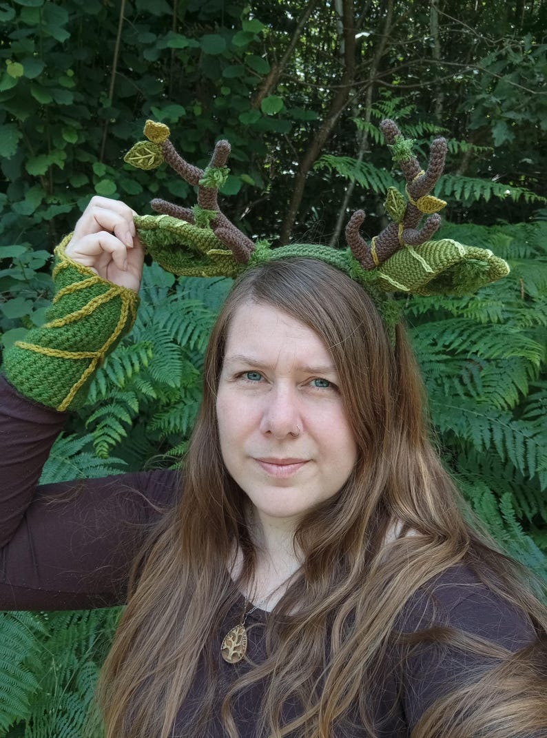 May include: A handmade crochet headband with deer antlers and matching arm cuffs. The antlers are brown with green leaves and the headband and cuffs are green with yellow accents. The person in the image is wearing a brown shirt.