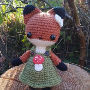 May include: A crocheted fox doll wearing a green dress with a mushroom applique and white flower on its head.