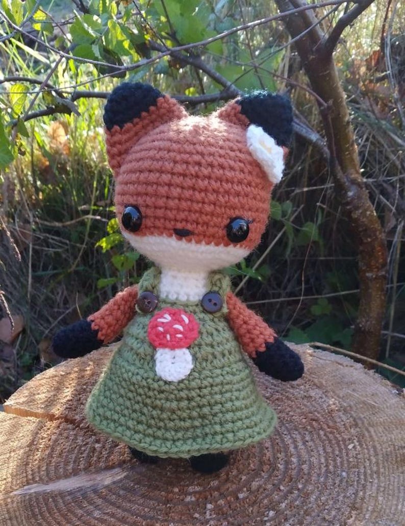 May include: A crocheted fox doll wearing a green dress with a mushroom applique and white flower on its head.