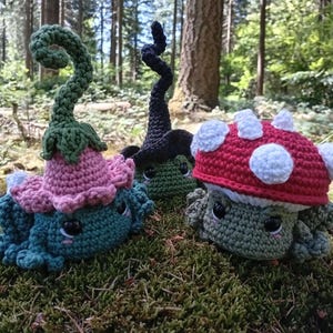 May include: Three crocheted plush figures with dark eyes and pink cheek accents. One is a teal creature with a pink flower-like hat and a tall green stem. Another is a dark green creature with a black pointed hat. The third is a light green creature with a red mushroom cap featuring white spots.