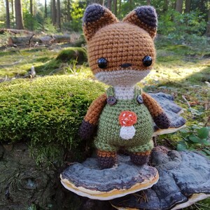 May include: A crocheted fox wearing green overalls with a mushroom design. The fox is standing on a mushroom-shaped wooden stump in a forest setting.