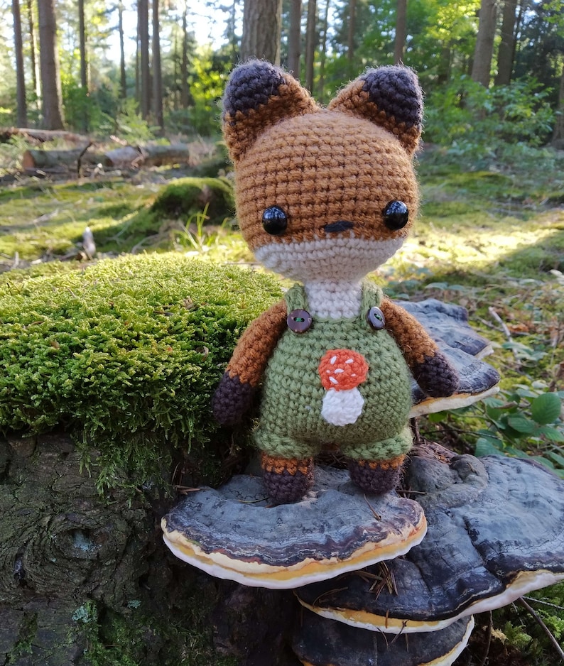 May include: A crocheted fox wearing green overalls with a mushroom design. The fox is standing on a mushroom-shaped wooden stump in a forest setting.