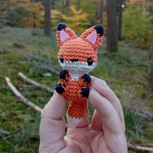 May include: A small, crocheted fox figurine. The fox is primarily orange with white, black, and pink accents. It has large, black eyes and is held in a person's hand against a blurred forest background.