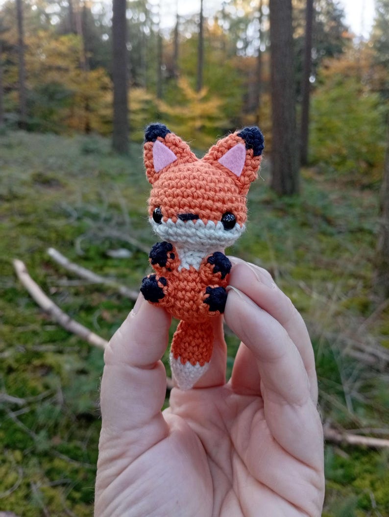 May include: A small, crocheted fox figurine. The fox is primarily orange with white, black, and pink accents. It has large, black eyes and is held in a person's hand against a blurred forest background.