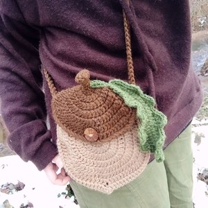 May include: A brown and tan crocheted acorn-shaped bag with a green crocheted leaf. The bag is worn as a crossbody bag.