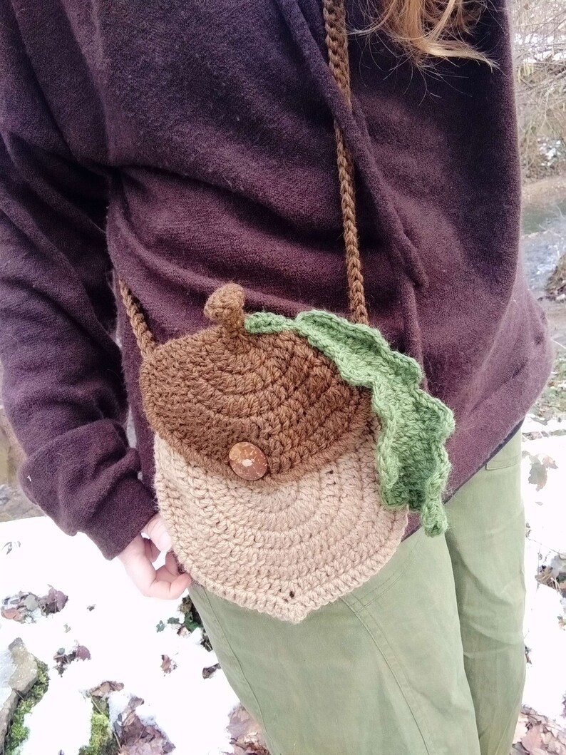 May include: A brown and tan crocheted acorn-shaped bag with a green crocheted leaf. The bag is worn as a crossbody bag.