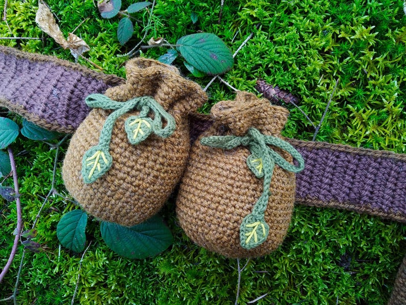 K&ouml;nnte beinhalten: Zwei braune H&auml;kelbeutel mit gr&uuml;nen Blattembroidery und braunen B&auml;ndern. Die Beutel liegen auf einem Bett aus gr&uuml;nem Moos.