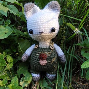May include: A white crocheted stuffed animal with gray ears and a green and brown crocheted overall. The overall has two buttons and a mushroom design on the front.