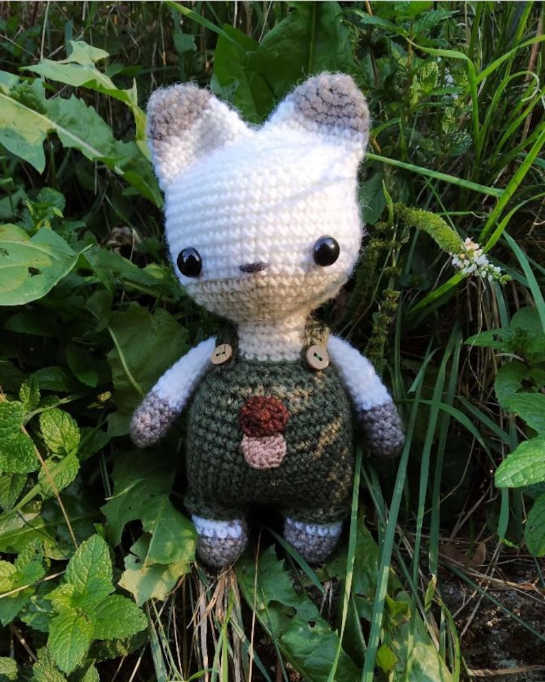 May include: A white crocheted stuffed animal with gray ears and a green and brown crocheted overall. The overall has two buttons and a mushroom design on the front.