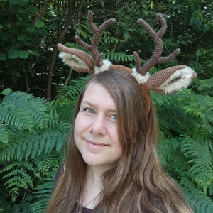 May include: A brown crocheted deer antler headband with faux fur-lined ears. The antlers are a darker shade of brown, and the headband is worn against a backdrop of green foliage. A whimsical accessory.