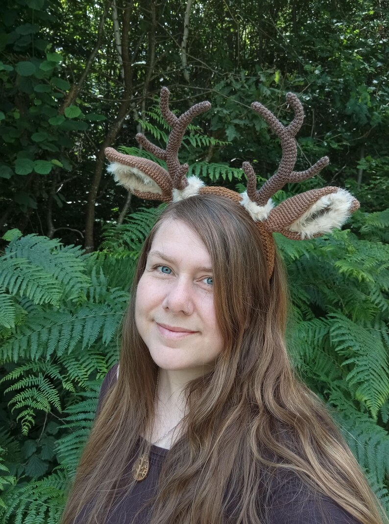 May include: A brown crocheted deer antler headband with faux fur-lined ears. The antlers are a darker shade of brown, and the headband is worn against a backdrop of green foliage. A whimsical accessory.