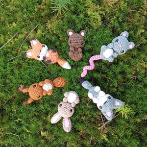 May include: A collection of handmade crochet woodland animal toys, including a fox, bear, mouse, squirrel, and rabbit. The toys are in various shades of brown, gray, and orange, with white and pink accents. They are arranged on a bed of green moss.