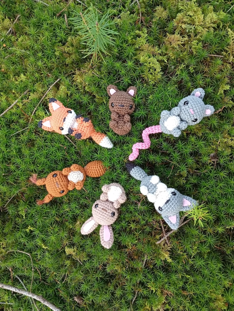 May include: A collection of handmade crochet woodland animal toys, including a fox, bear, mouse, squirrel, and rabbit. The toys are in various shades of brown, gray, and orange, with white and pink accents. They are arranged on a bed of green moss.
