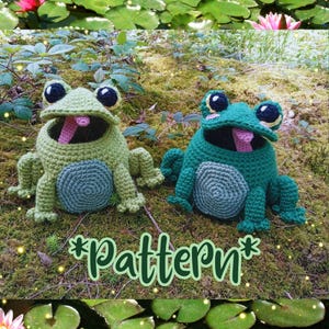 Peut inclure: Deux figurines de grenouilles au crochet, dans des tons de vert, avec de grands yeux noirs et jaunes et des langues roses. Les grenouilles ont un corps vert clair, un ventre vert foncé et sont posées sur une surface moussue. Le mot "Pattern" est écrit en vert.