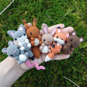 May include: A collection of handmade crochet woodland animal toys, including a gray wolf, brown squirrel, tan bunny, orange fox, and brown bear. Each animal has detailed features and is held in a hand against a mossy green background.