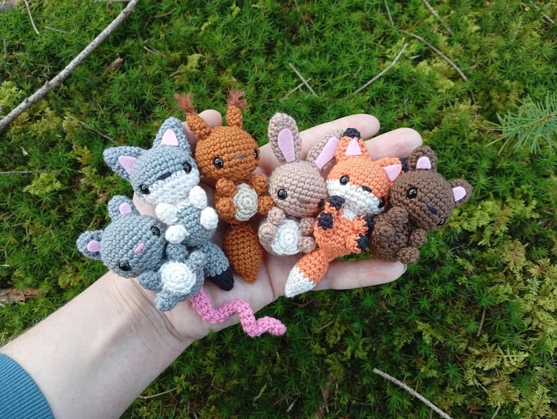 May include: A collection of handmade crochet woodland animal toys, including a gray wolf, brown squirrel, tan bunny, orange fox, and brown bear. Each animal has detailed features and is held in a hand against a mossy green background.