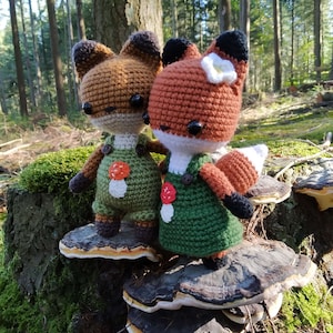May include: Two crocheted fox plushies wearing green overalls with mushroom accents, standing on a mushroom-shaped wooden stump in a forest setting.
