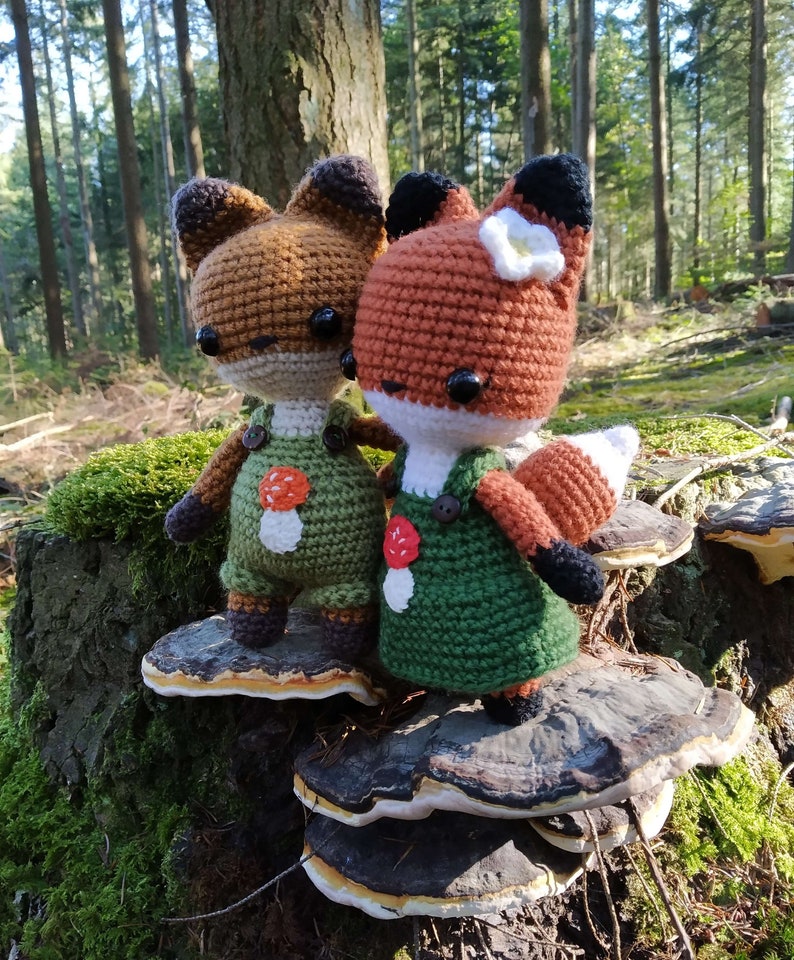 May include: Two crocheted fox plushies wearing green overalls with mushroom accents, standing on a mushroom-shaped wooden stump in a forest setting.