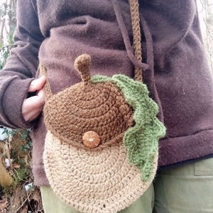 May include: A brown and beige crocheted acorn-shaped bag with a green crocheted leaf. The bag has a brown button and is worn as a crossbody bag.