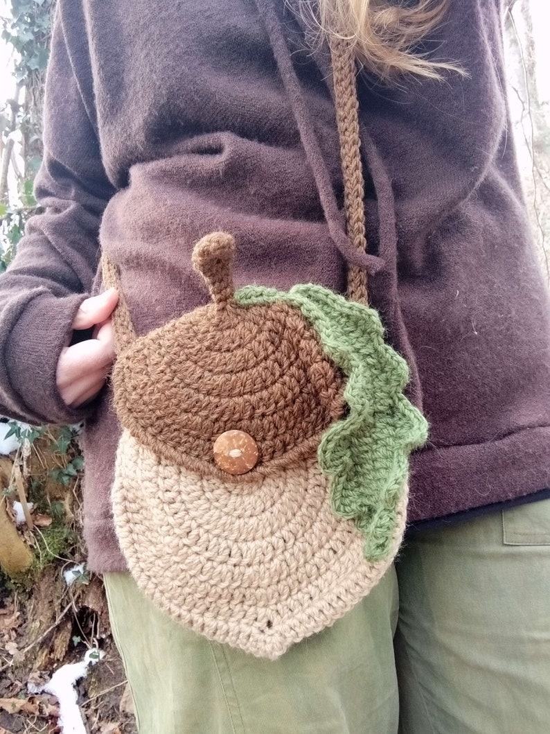 May include: A brown and beige crocheted acorn-shaped bag with a green crocheted leaf. The bag has a brown button and is worn as a crossbody bag.