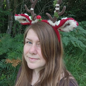 May include: A red and white crocheted reindeer antler headband with brown antlers, white fur trim, and decorative accents. The headband is worn by a person with long brown hair, set against a backdrop of green foliage.