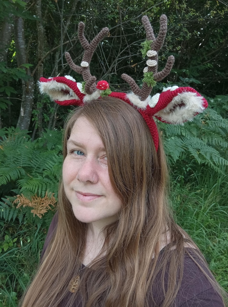May include: A red and white crocheted reindeer antler headband with brown antlers, white fur trim, and decorative accents. The headband is worn by a person with long brown hair, set against a backdrop of green foliage.