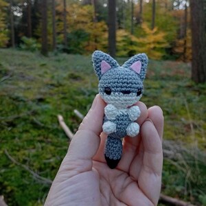 May include: A handmade crochet fox figurine in shades of gray, white, and black, with pink inner ears. The small, detailed fox is held in a hand against a blurred forest background, showcasing the craft's intricate details.